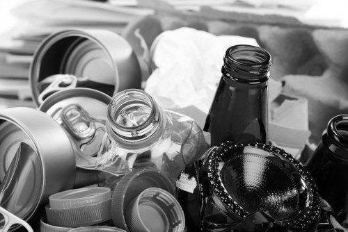 A collection of various types of rubbish including crumpled white paper, several empty and partially crushed black and transparent glass bottles, some metallic and plastic bottle caps, and a few discarded containers and packaging materials. The waste is scattered in an untidy manner, with the bottles lying on their sides and caps piled together. The objects are placed on a flat surface, possibly outdoors or in a storage area, with no visible background or environment. The image appears in black and white, emphasizing the textures and contrasts of the materials, which include smooth glass surfaces and rougher plastic and paper textures. This scene is representative of waste items that need removal and disposal, aligning with rubbish clearance services such as those offered by Office Clearance Islington.