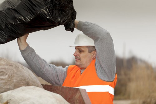 A male waste collection worker wearing an orange high-visibility vest and a white safety helmet is lifting a large black plastic rubbish bag. He appears to be outdoors, possibly on a construction site or a roadside area, with a background of blurred, natural landscape featuring bare trees and an overcast sky. The worker is focused on his task, holding the bag with both hands, preparing to load or transport it. Surrounding him are various objects, including a weathered stone or concrete block and some other unidentified materials, indicating a site designated for rubbish removal or clearance. The scene emphasizes the practical nature of waste collection work, aligned with services offered by companies like Office Clearance Islington, serving areas around Islington and London, where rubbish removal from commercial or residential properties is common. The textures of the bag contrast with the worker's smooth safety gear and the rough outdoor environment, underscoring the physical aspect of waste management duties performed in the local area.