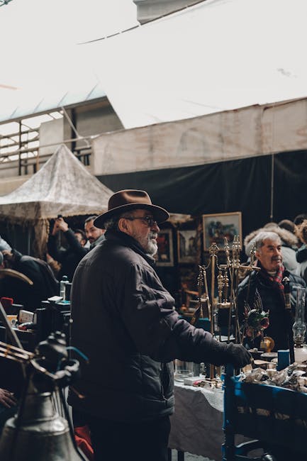 A man wearing a wide-brimmed hat, glasses, and a dark jacket is standing at a table filled with various decorative objects, including brass candlesticks, glassware, and small ornaments, at an indoor m