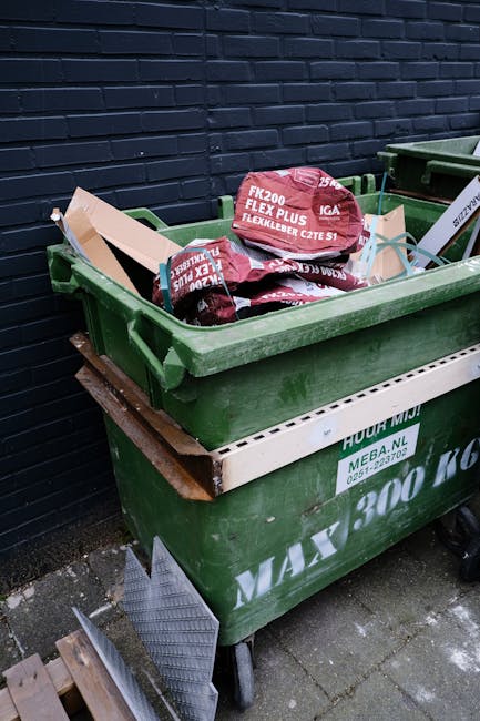 A large green wheeled waste skip positioned on a curb next to a dark brick wall, filled with several red and black bags of landscaping or gardening materials labeled 'FK200 FLEXY PLUS FLEXLEBER.' The