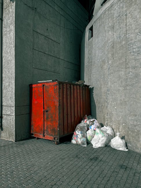 A large green wheeled waste skip positioned on a curb next to a dark brick wall, filled with several red and black bags of landscaping or gardening materials labeled 'FK200 FLEXY PLUS FLEXLEBER.' The